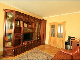 коричневая мебельная стенка с телевизором, часами, посудой за стеклом и полками с книгами в гостиной трехкомнатной квартиры