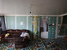 коричневый угловой мягкий диван на полу с ковровым покрытием с детским рисунком, шведская стенка у стены, с частичным ремонтом в детской спальне семейной дачи с недостроем
