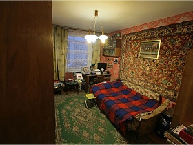 разложенный диван с сине красным покрывалом в клетку у стены с большим цветным ковром, книжные полки над письменным столом с настольной лампой, компьютером и бумагами в комнате с зеленым ковром на полу квартиры СССР 80-89 гг стиля
