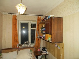 табурет у кровати с деревянными спинками, навесной шкаф с различными предметами на открытых полках спальной комнаты со светлыми обоями на стенах и желтыми шторами на окне двухкомнатной квартиры советского времени