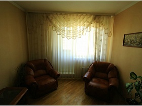 коричневые мягкие кресла у окна с балконом с нежной красиво оформленной бежевой гардиной в гостиной трехкомнатной квартиры