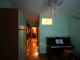 коричневый шкаф с голубой дверцей, стул у черного пианино с нотами в углу спальной комнаты с голубыми стенами и подвесной люстрой на белом потолке