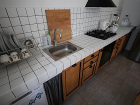 посуда в сушилке, электроприборы на столешнице из квадратной белой плитки над посудомоечной машинкой, коричневыми шкафчиками ,газовой духовкой в белой кухне зонированной комнаты скандинавской квартиры