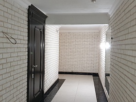 прямоугольное зеркало со светильником на кирпичной стене напротив черной входной двери в квартиру в светлом коридоре современного дома