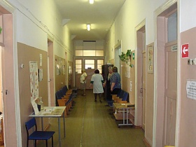 деревянные театральные кресла, банкетка, столики на деревянном полу длинного чистого коридора с освещением на белом потолке и розовыми панелями стен действующей больницы