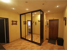 трехдверный коричневый шкаф-купе с зеркальными дверьми в углу просторной прихожей с деревянными масками, рамками, домофоном на бежевой стене прихожей и мягким пуфиком у входной двери простой московской квартиры