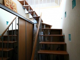 коричневый шкаф-купе с зеркальной дверцей под лестницей с деревянными ступенями и перилами, ведущая на второй этаж художественной дачи в сосновом лесу