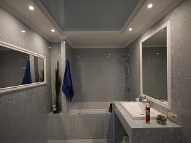 зеркала в белой рамке на стене, выложенной светло серой плиткой и над столиком с раковиной в ванной комнате с напольной стеклянной вазой у белой зашитой ванны и светильниками на натяжном потолке стильной дачи