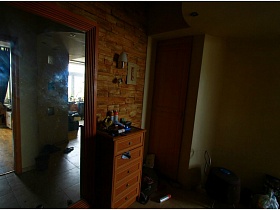 большое зеркало в деревянной рамке, коричневый комод у стены с плиткой под дикий камень у входной двери в прихожую двухкомнатной квартиры с видом на Москва-сити