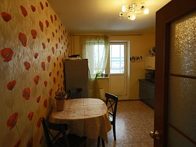 плетенная корзина с цветами на обеденном столе, серебристый холодильник, черно-белая мебельная стенка в светлой кухне с люстрой на белом потолке из открытой двери со стеклянными вставками