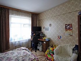 черное компьютерное кресло у стола с монитором, телевизором и бумагами, детские игрушки на полу, белое мягкое кресло у бежевой стены с фотографиями и перекидным календарем спальной комнаты просторной семейной квартиры в сталинском доме