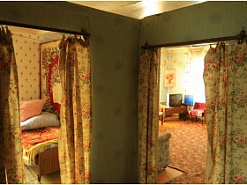 деревянная кровать с покрывалом и подушками, ковер на стене спальной комнаты и телевизор на тумбочке в углу гостиной через открытые дверные проемы с цветными шторами из прихожей жилого дома на селе