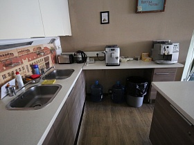 кофемашинки, электрочайник, тостер на светлой столешнице мебельной кухни с красочным оформлением фартука в офисном кафе