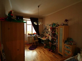 многочисленные детские игрушки на шкафу для одежды, подоконнике,письменном столе, полках мебельной стенки, комоде в детской комнате трехкомнатной квартиры с ораньжевой кухней