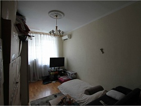 плоский телевизон на тумбе у окна с белой гпардиной в спальне простой квартиры на Садовом