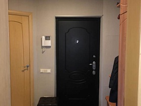мебельная стенка, черный пуфик, черная дорожка у входной двери прихожей с белым видеодомофоном на стене семейной квартиры
