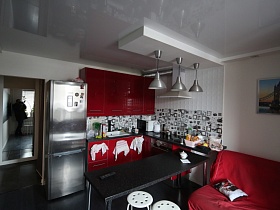 вишневая мебельная кухня с черной столешницей, черным обеденным столом, серебристым двухкамерным холодильником, белыми стульями на черном полу светлой комнаты современной квартиры