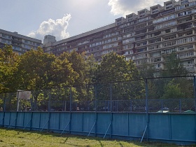 огромная спортивная площадка за голубым забором с высокой металлической сеткой на участке с подстриженной травой перед жилыми высотными домами в Чертаново
