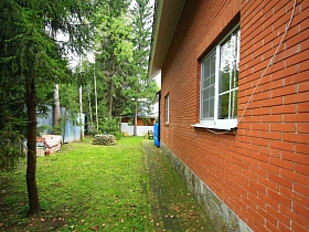 дорожка из плитки вокруг кирпичного двухэтажного дома с цоколем из дикого камня на зеленном участке за забором среди густого хвойного леса