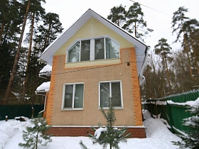 торец пустого двухэтажного современного дома под съем с треугольной крышей и отделкой на углах на зимнем участке в сосновом лесу