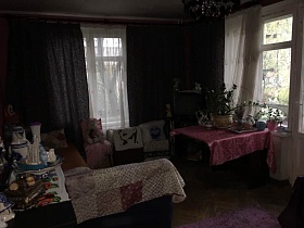 комнатные цветы на прямоугольном раскладном столе с сиреневой скатертью у окна с балконной дверью, стулья, комод и угловой диван у большого окна с темными шторами в гостиной с сиреневыми стенами квартиры в Останкино