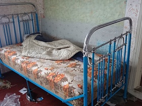 синяя железная кровать с одеялом на цветном матрасе в комнате с серыми обоями, окрашенным деревянным полом старого дома заброшенной деревни