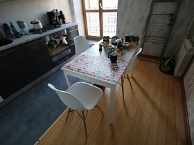 белые и серые дверцы мебельной стенки со светло серой столешницей под мрамор на полу с крупной серой плиткой, белые стулья со спинкой вокруг белого обеденного стола на полу с ламинатом стильной кухни трехкомнатной квартиры