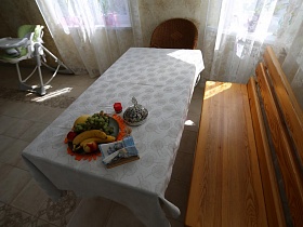 длинная деревянная скамейка со спинкой у обеденного стола с белой скатертью, коричневым креслом у окна с белой гардиной кухни семейной уютной дачи с частичным недостроем