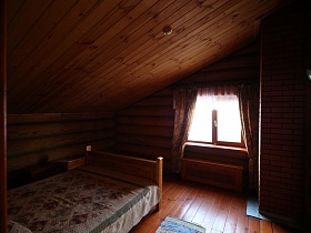 небольшой коврик у деревянной кровати с цветным покрывалом в мансарде со шторами на окне бревенчатого дома