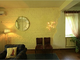квадратная подушка на большом кресле, два стула и торшер у желтой стены под кирпич с круглым окном на втором этаже современного дома под съем