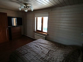 люстра на потолке с белыми цветочными плафонами, плоский телевизор на коричневом комоде у стены за дверью в светлой спальне деревянного дома