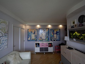 разноцветные дверцы шкафчика на колесах у светло серой стены с подсветкой и большими картинами в просторной гостиной стильной трехкомнатной квартиры в Икеа стиле