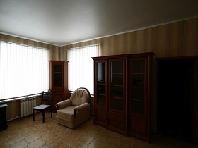 коричневые шкафы с пустыми полками за стеклом, мягкое бежевое кресло у окон с белыми жалюзи гостиной двухэтажного современного дома под съем