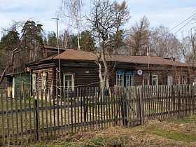 общий вид неокрашенного бревенчатого длинного многоквартирного дома во дворе за штакетным забором в Акуловке на торфоразработках