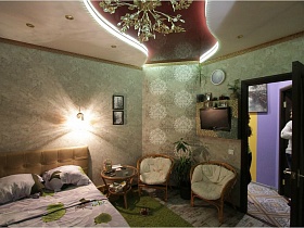 телевизор над плетенными креслами, комнатный цветок и круглы журнальный столик со стеклянной поверхностью в спальне с фигурным потолком в квартире с выходом на крышу магазина