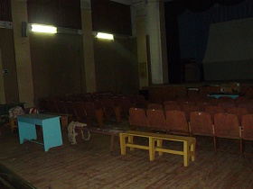 деревянные скамейки, ярко голубой стол за рядами секционных кресел на деревянном полу, требующем покраски актового зала старого клуба СССР