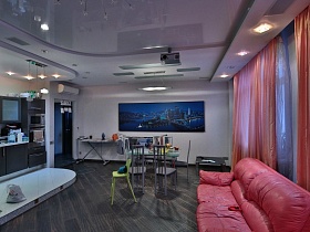 серая кухня на подиуме, квадратный журнальный столик, обеденный стол со стульями, черный телевизор на стене фиолетовой зонированной гостиной  с красным диваном и фиолетовым натяжным потолком