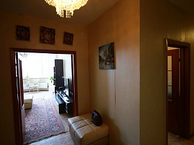 картины на бежевой стене над открытой дверью в просторную светлую гостиную, мягкий белый пуфик у двери в большом коридоре простой современной трехкомнатной квартиры на 6 этаже