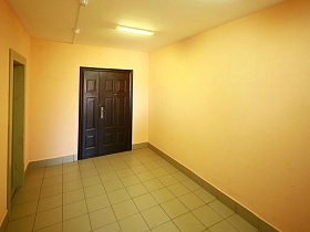 песочного цвета стены лифтового холла с квадратной плиткой на полу перед лифтами и входной дверью квартиры в многоэтажном доме