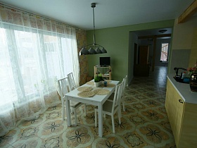телевизор на открытой деревянной полочке, белый обеденный стол с салфетками и белые стулья со спинкой на полу с цветной плиткой в красивой кухне с бежевой мебелью элитного недостроенного дома