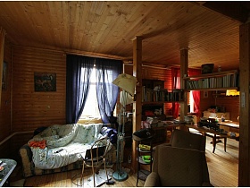 светлое одеяло на мягком диване у окна с синими шторами, ноутбук на стуле и торшер в гостиной деревянной дачи музыканта