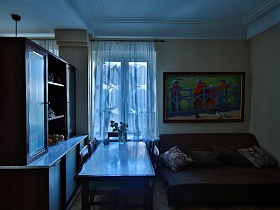 яркая картина с изображением детей над коричневым диваном с подушками и обеденный стол со стульями у окна гостиной квартиры художника