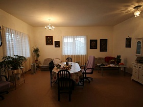 сиреневое кресло и коричневые стулья со спинками вокруг обеденного стола с цветной скатертью, люстры на потолке светлой столовой гостевого двухэтажного дома с придорожным кафе