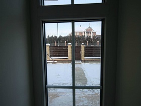 вид из окна на коричневый забор с колоннами вокруг зимнего участка с расчищенной дорожкой