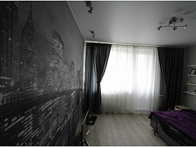 люстра и светильники в стиле хай-тек на белом потолке гостиной с фотообоями на стене, угловым разложенным диваном с фиолетовым покрывалом, темно серыми шторами и белой гардиной на окне евро однушки