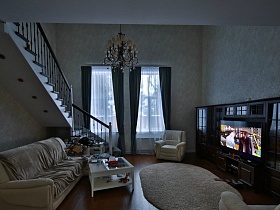 коричневая мебельная стенка с телевизором, белая мягкая мебель и стеклянный журнальный столик с светлой гостиной с лестницей на второй этаж добротного кирпичного дома