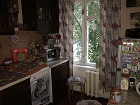 темно-коричневая мебельная стенка с белой столешницей, стиральная машинка между шкафами в кухне с цветными сиреневыми шторами квартиры в Останкино