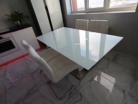 белая поверхность стеклянного стола, белые мягкие стулья со спинкой на металлических ножках на полу кухни с серой под мрамор плиткой
