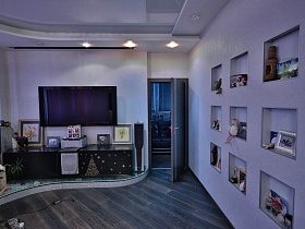 большой черный плоский телевизор, картины, фотографии, сувениры на поверхности двухцветной мебельной низкой стенки на подиуме в гостиной с сиреневыми стенами и серым полом квартиры студии молодежной семьи