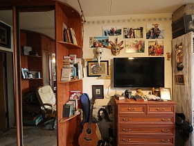 гитары у боковых полок с книгами коричневого шкафа-купе, коричневый комод у светлой стены с плоским телевизором, яркими фотографиями, различными масками в спальной комнате трехкомнатной квартиры